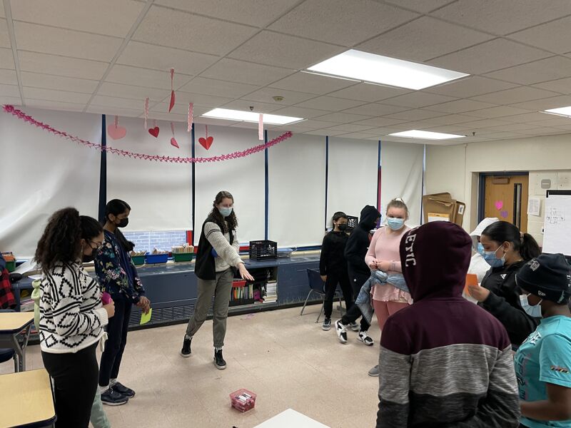 The image shows a group of students in a classroom. Some students are wearing masks. There are decorations hanging from the ceiling. It looks like they are doing some kind of activity. The room is well-lit.
